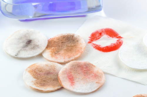 close up of makeup pads covered in various cosmetic products wiped from a face, with a purple bottle of cleanser in the back on a white background